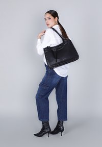 Donna in camicia bianca, jeans blu, stivali neri con tacco alto, che porta a spalla una grande borsa in pelle nera con bordi borchiati.