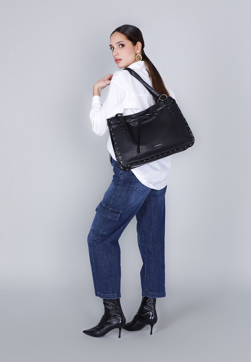 Donna in camicia bianca, jeans blu, stivali neri con tacco alto, che porta a spalla una grande borsa in pelle nera con bordi borchiati.