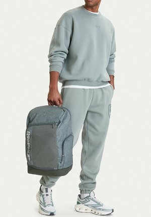 Uomo con felpa e pantaloni della tuta grigi coordinati, scarpe da ginnastica bianche, che tiene uno zaino Reebok grigio per la maniglia superiore.