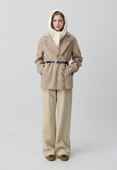 Femme portant un manteau beige à franges avec une ceinture noire, une écharpe à capuche en tricot crème, un pantalon large beige et des chaussures brunes sur un fond uni.