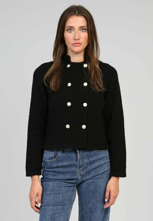 Veste noire texturée au design court, avec un col montant et des accents de boutons en argent à double boutonnage. Associée à un jean bleu.