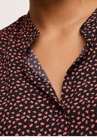 Blouse noire avec col, petits motifs de feuilles roses et texture légère. Des boutons sont présents le long du plastron.