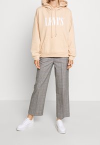 Felpa beige con il logo bianco "LEVI'S" e cordini, abbinata a pantaloni a gamba larga grigi a quadri e sneakers bianche.