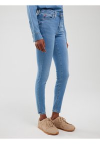 Isik kannab helesiniseid kitsaid teksapükse, sobivat teksasärki ja beeže pitsilise nöörimisega spordijalatsit, seistes ühevärvilise valge tausta ees.