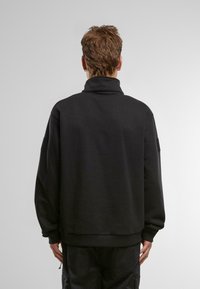 Črna sweatshirt s visokim ovratnikom in spuščeni rameni, izdelana iz gladkega materiala. Ima pravokotni znak na levi roki.