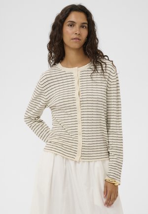 Woman wearing a cream cardigan with black horizontal stripes and a white skirt, posing with one hand on her hip against a plain background.
