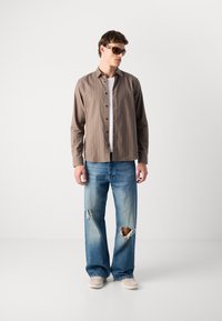 Camicia button-up a righe marrone chiaro, t-shirt bianca sotto, jeans larghi blu strappati e scarpe beige senza lacci. Inclusi occhiali da sole.