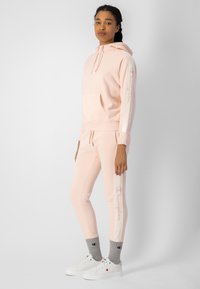 Sudadera rosa clara con bolsillo frontal y detalles blancos en las mangas, combinada con leggins a juego. Calcetines grises y zapatillas blancas completan el conjunto.