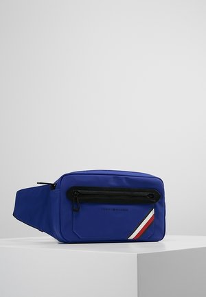 Blå Tommy Hilfiger bæltetaske med sort lynlåslomme, rød-hvid-blå diagonalstribe og justerbar rem på hvid overflade.
