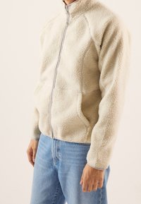 Fleece jacket in cream color with a textured exterior, full zip closure, and stand collar. Features gray accents and side pockets.
