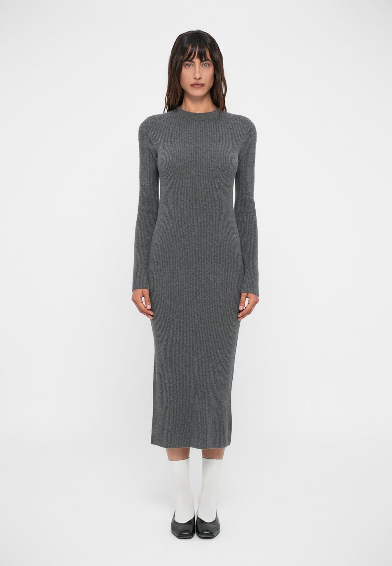 Femme se tenant contre un fond uni, portant une robe midi grise à manches longues ajustée, des chaussettes blanches et des chaussures noires.