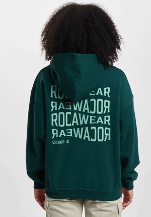 Felpa verde scuro realizzata in morbido tessuto, caratterizzata da un grande testo bianco sul retro che mostra "ROCAWEAR EST.1999" in motivi sovrapposti.