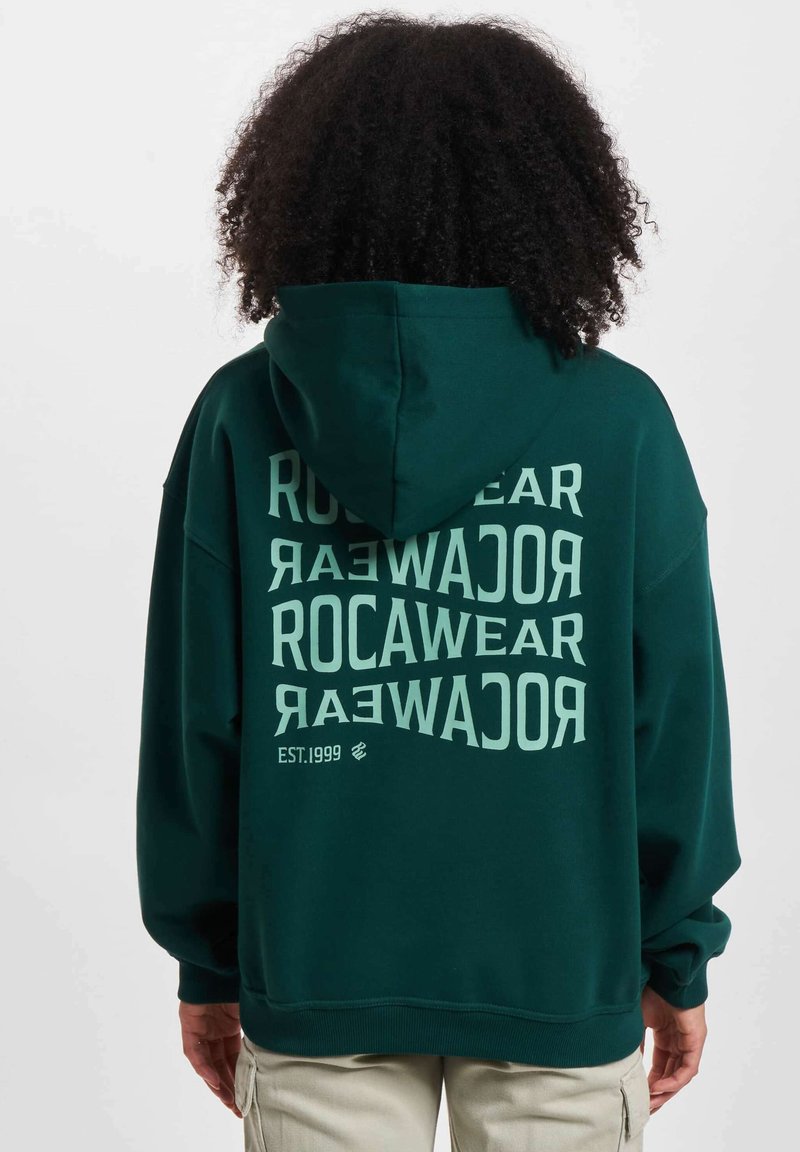 Dark green hoodie made of soft fabric, featuring large white text on the back, displaying "ROCAWEAR EST.1999" in overlapping patterns.
