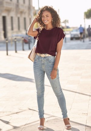Bordeauxrotes, geripptes Kurzarmoberteil, hellblaue Skinny-Jeans, beiger Gürtel und weiße High Heels. Steht im Freien und lächelt.