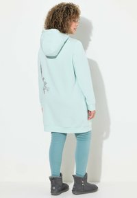 Abito felpa verde menta con cappuccio, tasche laterali e dettaglio floreale sulla parte posteriore, abbinato a leggings teal e stivali alla caviglia grigi.