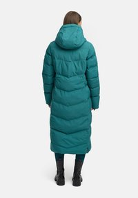 Tamsi zaļa garā pūkainā jaka ar kapuci, ar chevron šuvju dekoriem, sānu rāvējslēdzēju un brīvu piegriezumu. Valkāta virs tumšām legingām un melniem zābakiem.