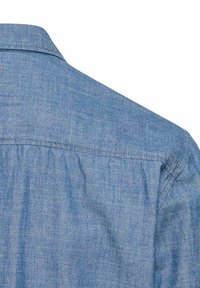 Niebieska koszula chambray z wyraźnym kołnierzem, przeszyciem na karczku z tyłu oraz subtelną fakturą. Tkanina wydaje się lekka i lekko szczotkowana.