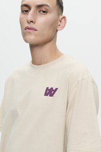 Jauns vīrietis ar īsiem tumšiem matiem valkā bēšīgu T-kreklu ar tumši violetu W logotipu uz kreisā krūtīm, nofotografēts pret vienkāršu gaišu fona.