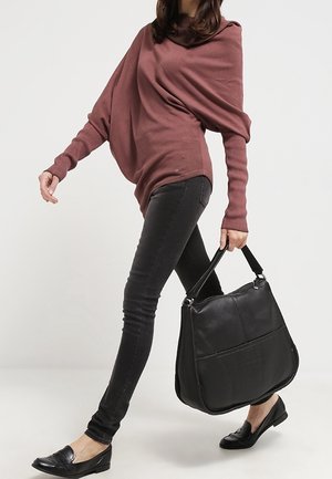 Frau, die einen lockeren puderrosa Pullover und schwarze Skinny-Jeans trägt, geht und eine große schwarze Ledertasche trägt und schwarze Loafers anhat.