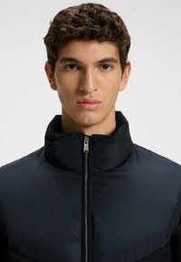 Zwarte zip-up jas met een hoge kraag, gemaakt van glanzende stof. Voorzien van een zilveren rits en een soepele textuur, met een naadloos ontwerp.