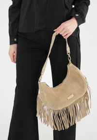 Beige suède schoudertas met franjes, gouden hardware en een rits-sluiting. De schouderband is verstelbaar voor extra comfort.