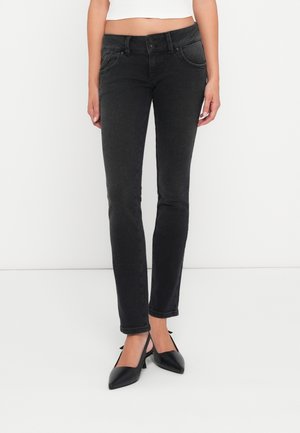 Femme portant un jean noir ajusté et des chaussures noires à talons bas et bouts pointus, debout devant un fond blanc uni.