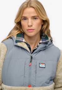 Graue isolierte Weste mit beigen, flauschigen Ärmeln, ausgestattet mit einem Reißverschluss, einem Karottenhemdkragen und einem Logopatch nahe dem vorderen Reißverschluss.