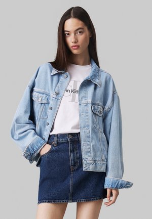 Chaqueta de mezclilla azul claro con mangas largas y cierres de botones, llevada sobre una camiseta blanca con un logotipo gris, combinada con una falda de mezclilla oscura.
