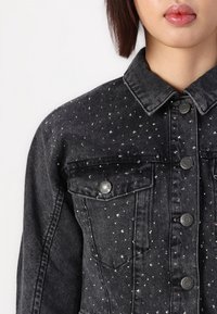Czarna kurtka dżinsowa z srebrnymi cekinami, klasyczny kołnierz, przednie kieszenie i metalowe zapięcia. Teksturowany materiał.