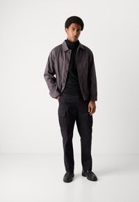 Chaqueta de mezclilla gris oscuro, suéter de cuello alto negro, pantalones cargo y zapatos negros. Ajuste casual con cierre de botones, tela texturizada y bolsillos utilitarios.