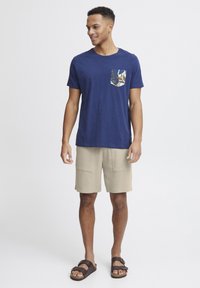 Tricou bleu marin cu mâneci scurte și buzunar cu model; pantaloni scurți bej texturat cu două buzunare mari în față; sandale maronii.