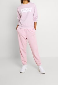 Sweat-shirt Levi's rose pâle avec logo blanc, assorti d'un pantalon de jogging assorti. Le tissu semble doux et confortable ; poignets élastiques aux chevilles.