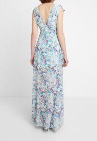 Femme portant une robe sans manches, longue jusqu'au sol, de couleur bleu pastel avec des motifs floraux et un dos en V profond, se tenant devant un fond blanc uni.