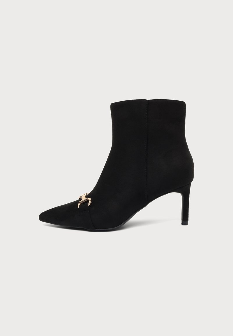 Bottine en daim noire à bout pointu avec talon haut fin et boucle décorative dorée sur le côté, sur fond uni.