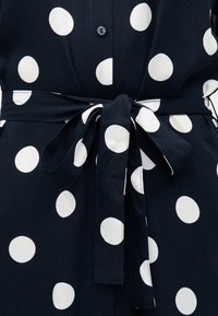 Ύφασμα σε ναυτικό μπλε με μεγάλα λευκά πουά, με ζώνη δεμένη στη μέση και ένα μικρό μαύρο κουμπί στο κέντρο.