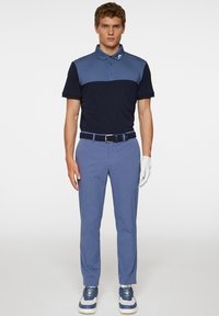 Marineblaues und himmelblaues Polohemd mit kurzen Ärmeln, kombiniert mit hellblauen Hosen und weißen Sneakers. Mit Logo am Kragen und geflochtenem Gürtel.