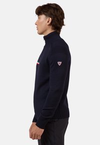 Maglione blu navy a maglia con colletto alto, decorato con una striscia orizzontale rossa e bianca e un patch con logo sulla manica sinistra.