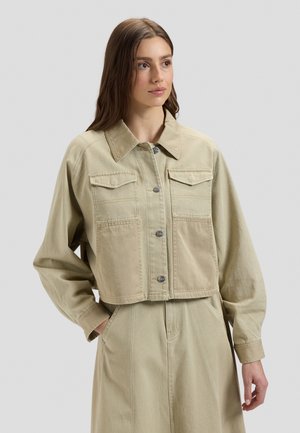 Jonge vrouw met lang bruin haar, gekleed in een beige, kort jasje met knopen en een bijpassende high-waisted rok, staand tegen een effen achtergrond.
