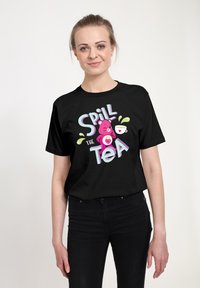 Camiseta de algodón negra con un gráfico divertido que muestra un oso rosa y el texto "Spill the Tea" en letras coloridas, presentado sobre un fondo neutro.