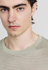 Primo piano di un uomo con capelli scuri, pelle chiara, occhi blu, che indossa un maglione lavorato a maglia verde chiaro con colletto rotondo.