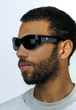 Uomo con capelli corti e ricci e barba, che indossa occhiali da sole neri avvolgenti e una maglietta grigio chiaro con scollo tondo, rivolto verso sinistra.
