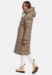Lange, taupe pufferjas met capuchon, gevoerd ontwerp, voorzakken en ritsen. Draagt over witte broeken en beige laarzen. Zwarte accenten zichtbaar.