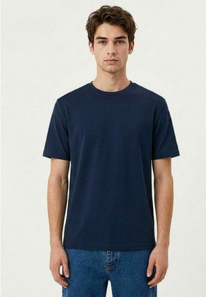 Jonge man met kort bruin haar, die een effen donkerblauw T-shirt met korte mouwen en blauwe spijkerbroek draagt, staand tegen een lichte achtergrond.
