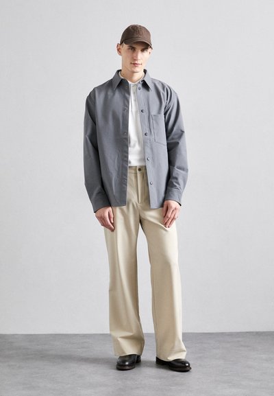 Gray button-up shirt over a white t-shirt, paired with light beige trousers and dark shoes, accessorized with a brown cap.