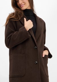 Brauner Wollmischungsmantel mit einem Reverskragen, zwei vorderen Taschen und großen schwarzen Knöpfen. Weiche Textur, lockere Passform und lange Ärmel.