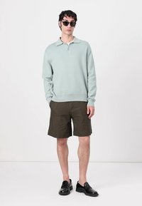 Light blue ribbed sweater with a polo collar, paired with olive green shorts. Model wears dark sunglasses and black slip-on shoes.