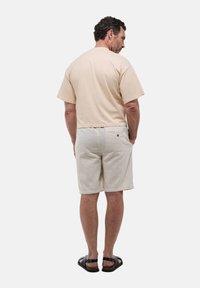 T-shirt en coton beige avec des manches courtes, associé à un short tissé de couleur claire avec une poche arrière unique et un détail de bouton. Sandales noires.