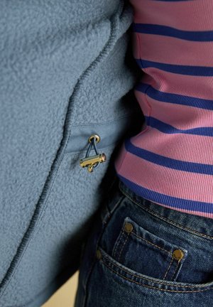Close-up of two people standing side by side, one wearing a blue fleece jacket with a gold toggle, the other in a pink and blue striped top with denim jeans.