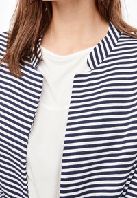Veste blazer rayée avec des lignes horizontales bleu marine et blanches, tissu léger, devant ouvert et col étroit portée sur un haut blanc.