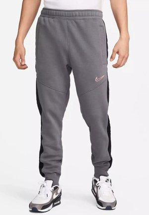 Mężczyzna ubrany w szare spodnie dresowe Nike z czarnymi bocznymi panelami oraz białe buty sportowe Nike na białym tle.